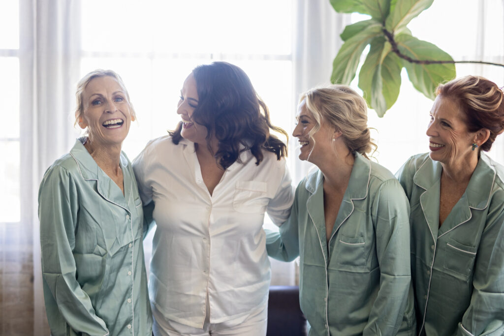 bride laughing with bridesmaid and moms in matching PJs getting ready