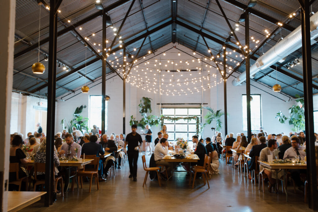 Wide view of a wedding reception inside a large, modern venue with string lights and guests seated at tables.