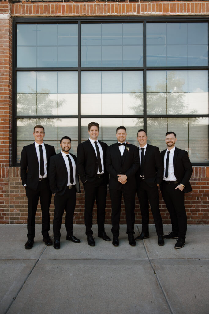 Groom and groomsmen standing in a line, smiling outside a brick and glass building.