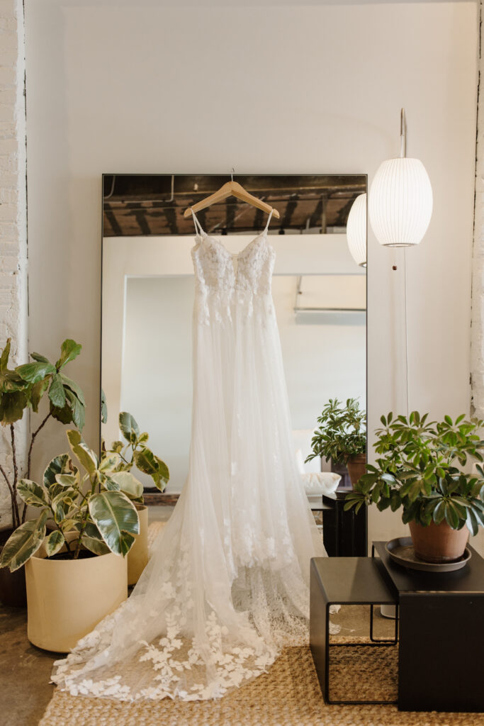 Wedding dress hanging on a mirror between potted plants in a softly lit room.