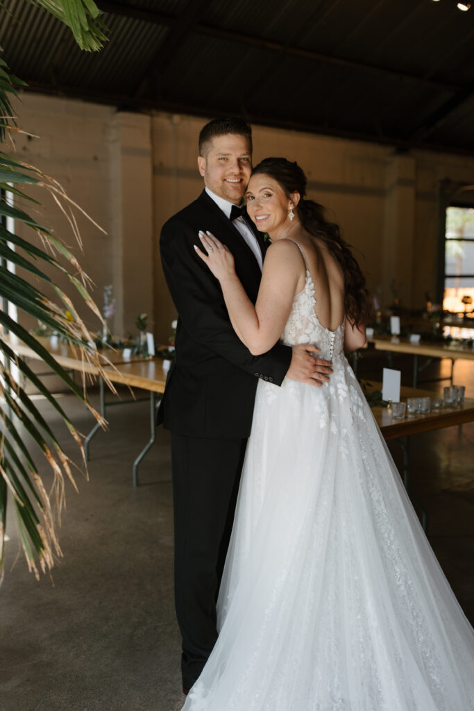Bride and groom embracing indoors near decorative plants and soft lighting.