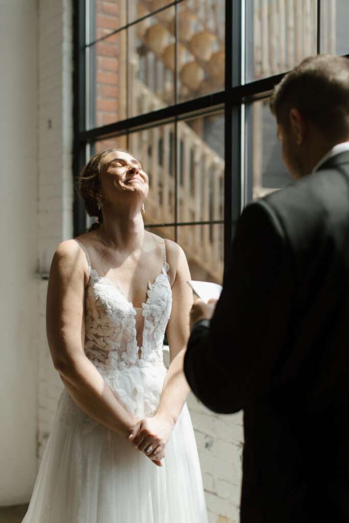 Bride smiling joyfully while holding hands with groom near a sunlit brick building.