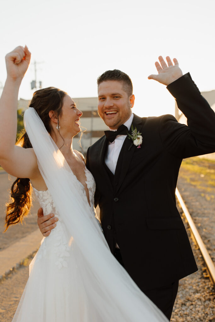 Bride and groom joyfully raising their arms in celebration outdoors, smiling at each other.