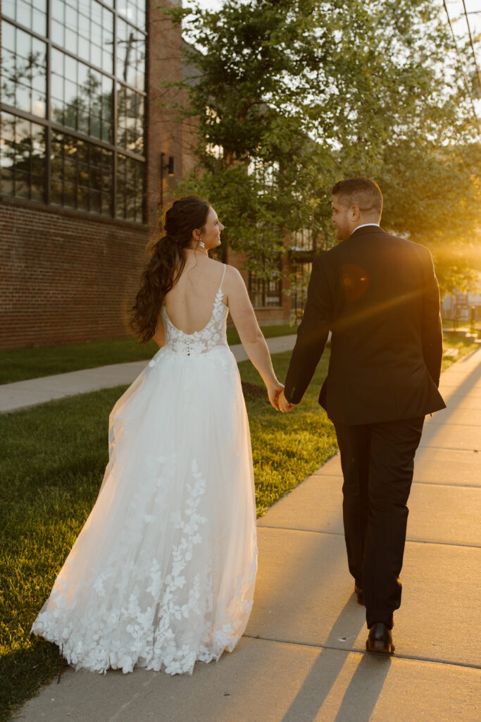 Bride and groom walking hand in hand during golden hour on a paved path.