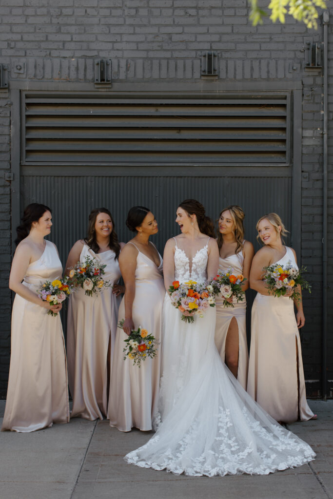 Bride and bridesmaids smiling together in front of a dark industrial wall, holding colorful bouquets.