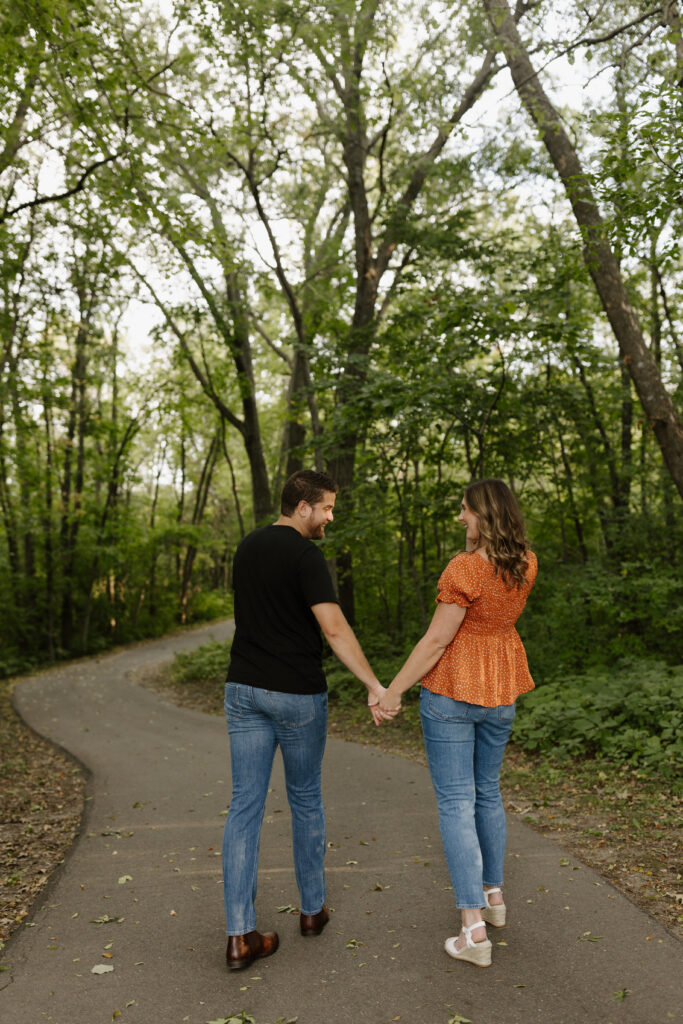 Engagement photo of a couple holding hands and walking on a wooded path.