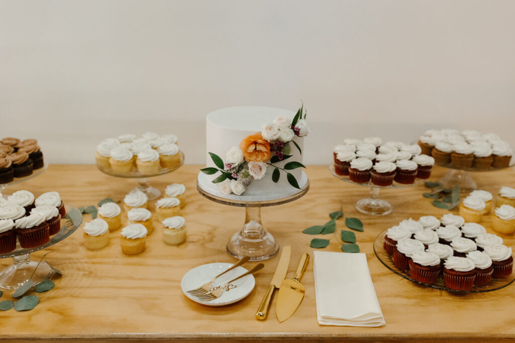 Minimalist dessert table with a white wedding cake, cupcakes, and gold cutlery.