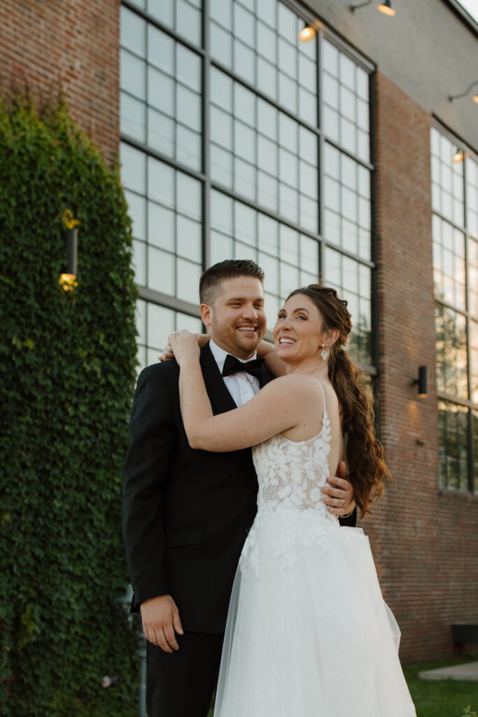 Bride and groom hugging outside near a building with tall windows and green ivy.