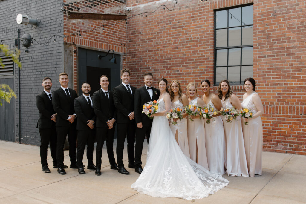 Full wedding party posing outdoors against a brick wall, bride in the center holding bouquet.