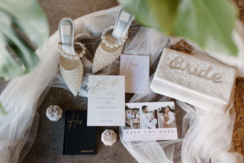 Flat lay of wedding accessories including invitation suite, rings, perfume, and shoes.