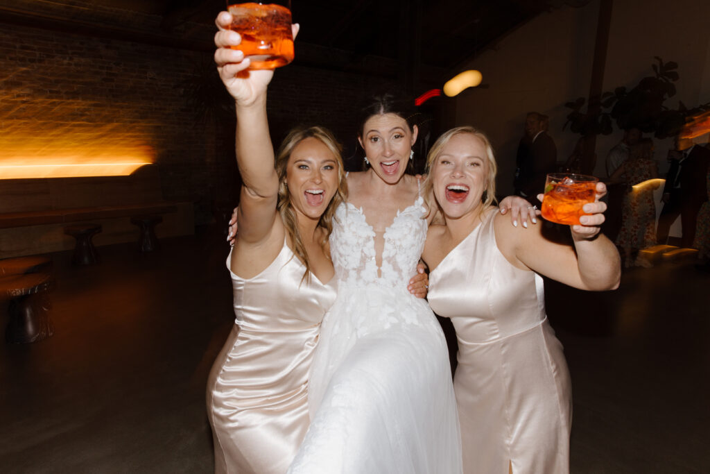 Bride and bridesmaids in satin dresses celebrating with raised cocktails.
