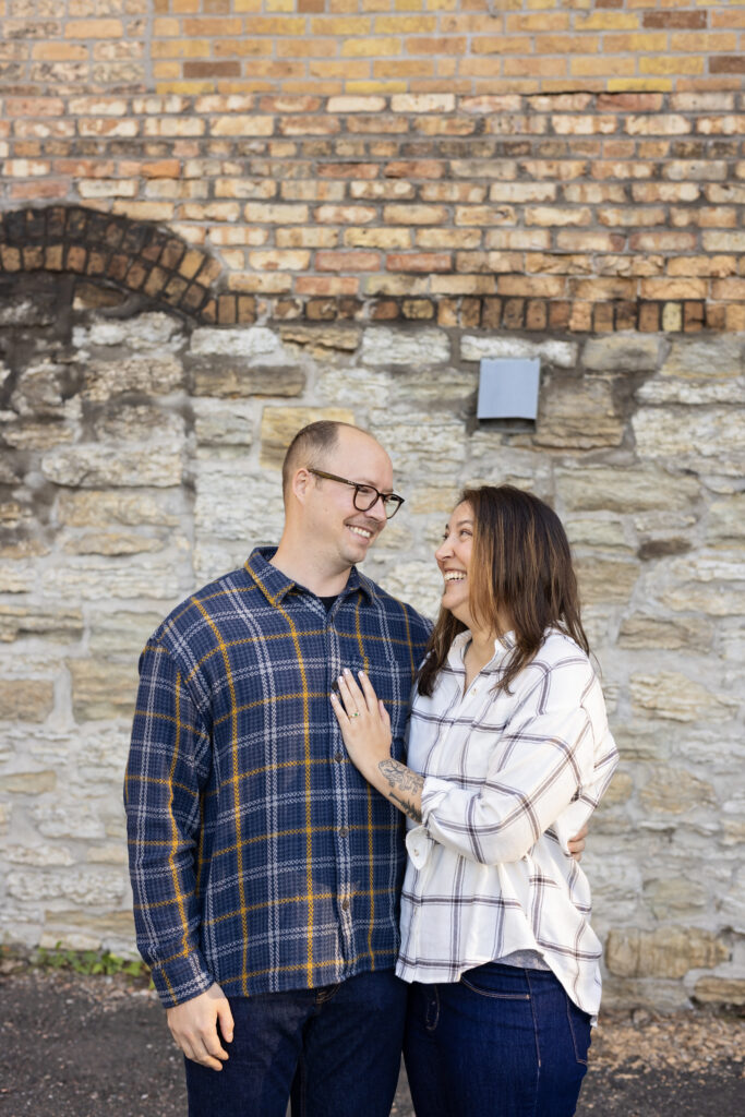 A couple stands close together, smiling and laughing, in front of a rustic brick and stone wall.
