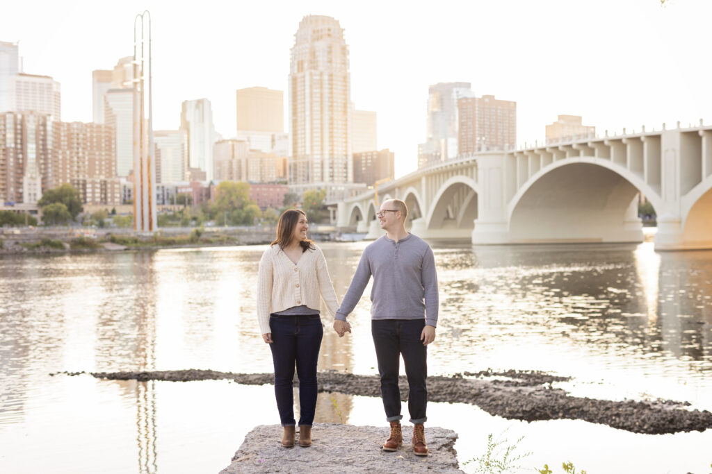 The couple holds hands by a riverside with the Minneapolis skyline and bridge in the background, bathed in golden hour light.