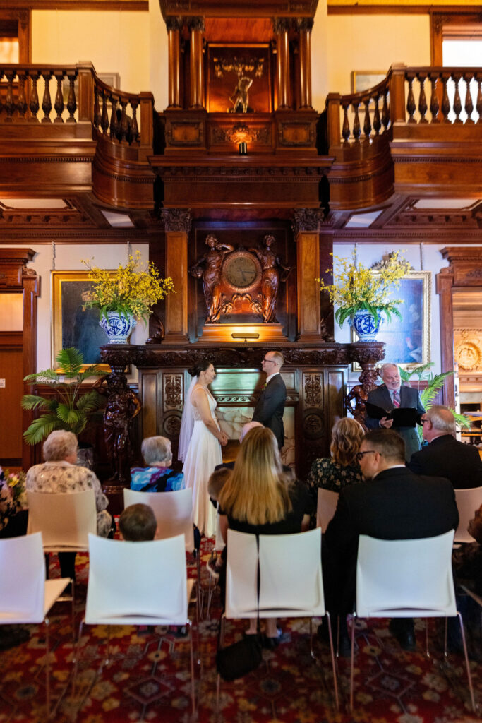 Bride and groom stand together facing the officiant during their ceremony, surrounded by guests in an elegant, historic room.