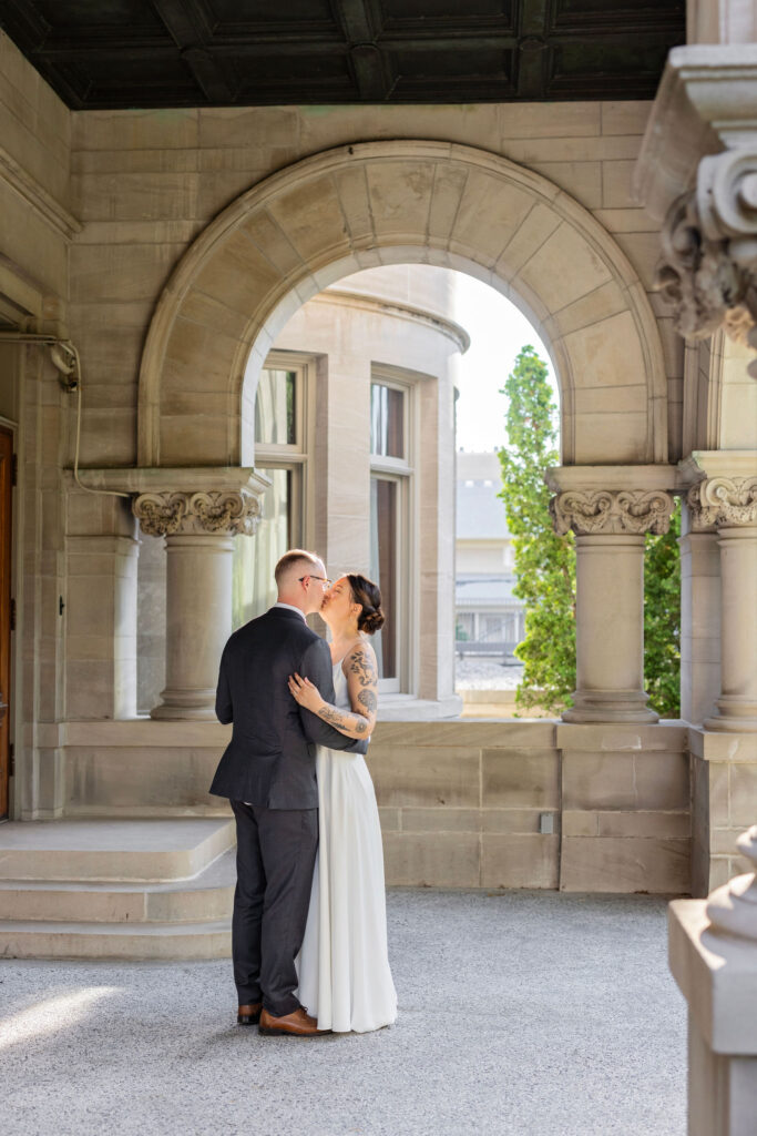 Bride and groom share a private kiss beneath a stone archway of a historic building, framed by natural light.