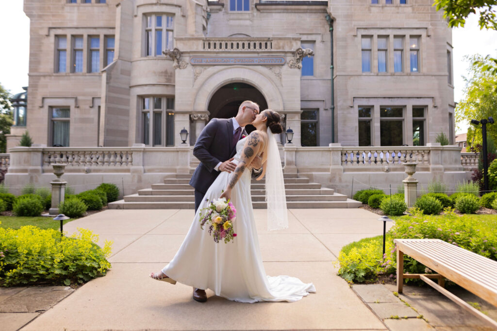 A bride and groom share a romantic kiss in front of the grand American Swedish Institute, with the bride holding a colorful bouquet.