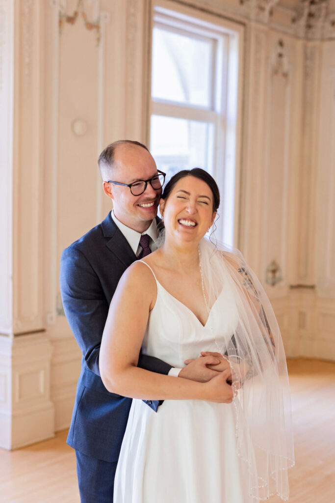 Groom embraces the bride from behind as she laughs, both standing in a softly lit, elegant indoor space with tall windows.