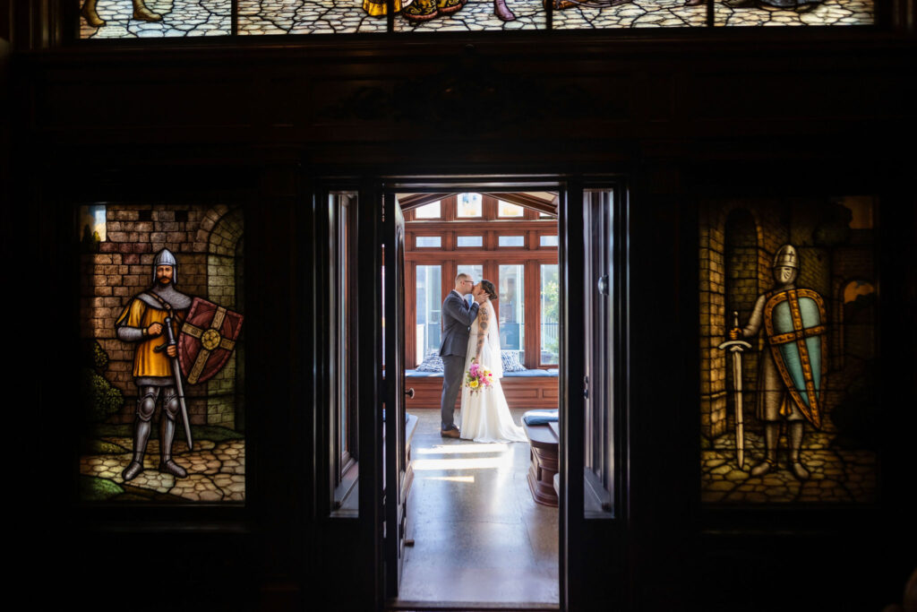 A bride and groom share an intimate moment in a sunlit room framed by dark wood doors and medieval stained glass windows featuring knights.