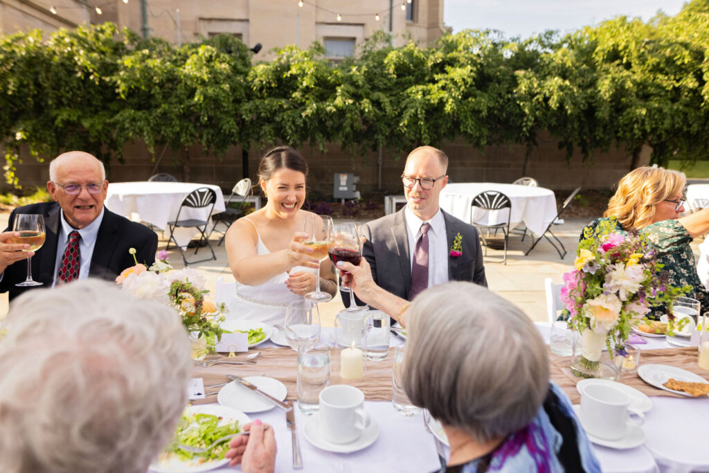 Bride laughs while clinking glasses with guests during an outdoor dinner toast, groom smiling beside her.