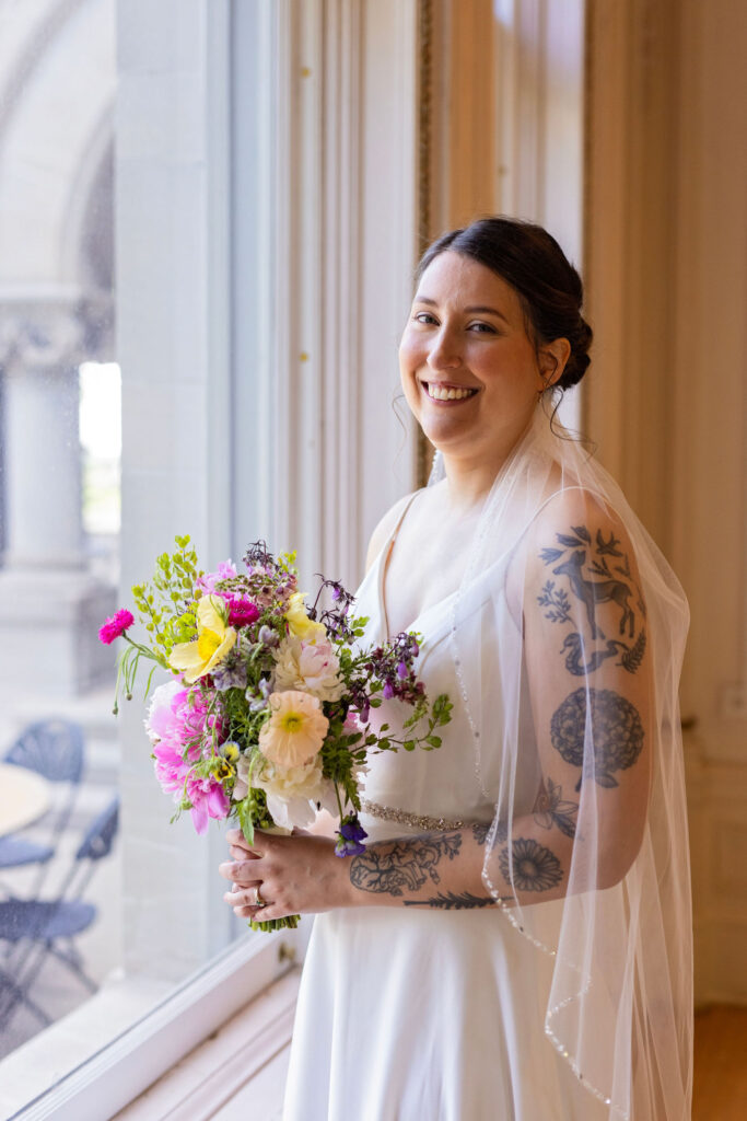 Bride smiles while holding a colorful bouquet, standing by a window in an elegant room, her tattooed arm visible beneath her veil.