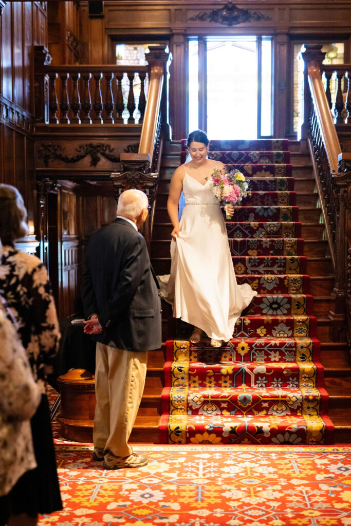 Bride walks gracefully down an ornate staircase in a grand wood-paneled hall as guests look on.