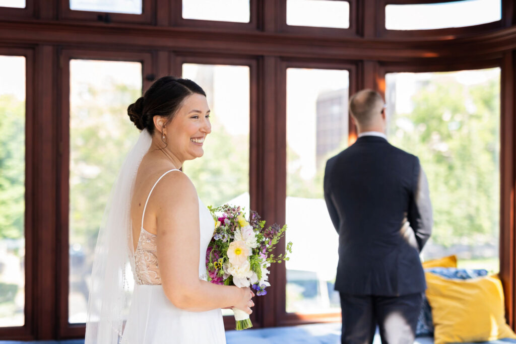 Bride approaches the groom from behind, smiling, holding her bouquet, with soft natural light streaming into a wooden-windowed room.