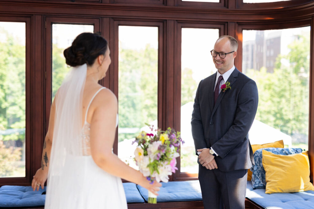 Bride and groom share a joyful first look, smiling at each other in a cozy sunroom filled with bright cushions and window light.