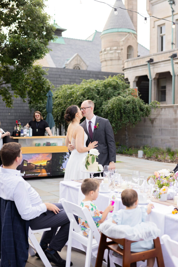 Bride and groom share a sweet kiss by the cake table while guests watch from nearby tables.