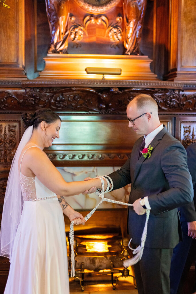 Bride and groom smile during a handfasting ceremony, wrapping cords around their joined hands in front of a carved fireplace.
