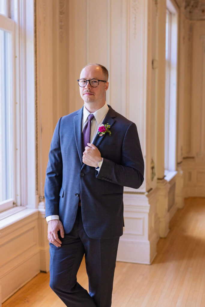 Groom in a navy suit stands confidently near a large window with ornate molding, adjusting his boutonniere.