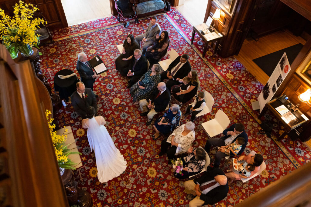 Overhead view of a micro-wedding ceremony, with the bride and groom standing at the front and guests seated closely on a patterned red carpet.