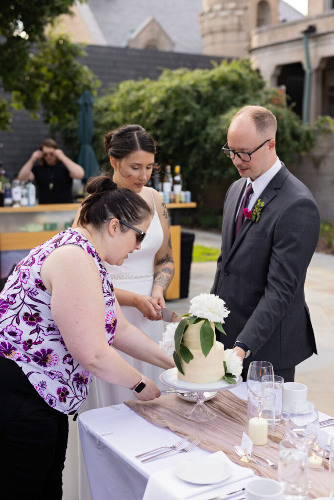 Wedding planner helps guide the couple in slicing their cake while they watch closely.