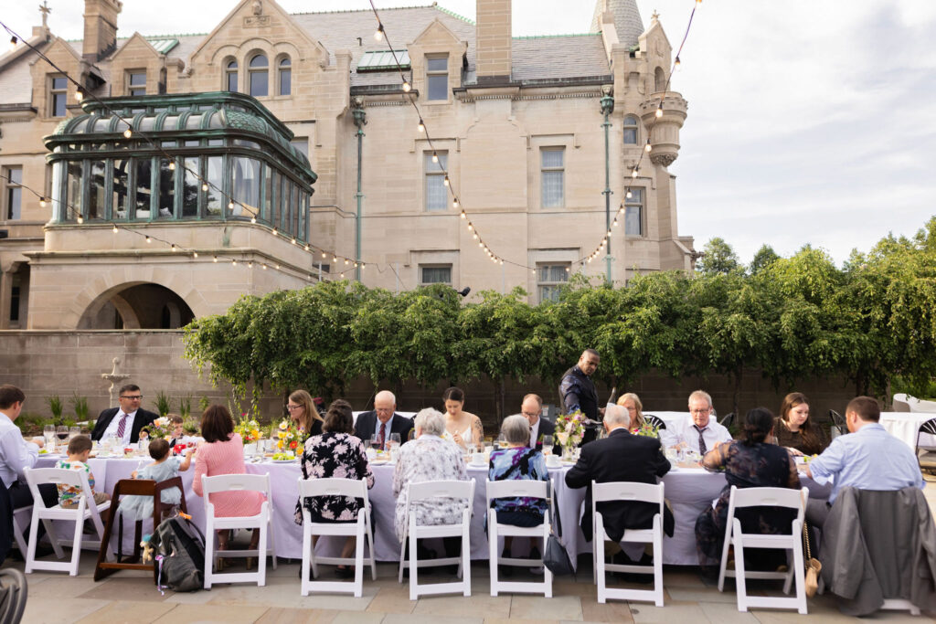 Wedding guests enjoy dinner together at a long outdoor reception table beneath string lights, with the American Swedish Institute in the background.