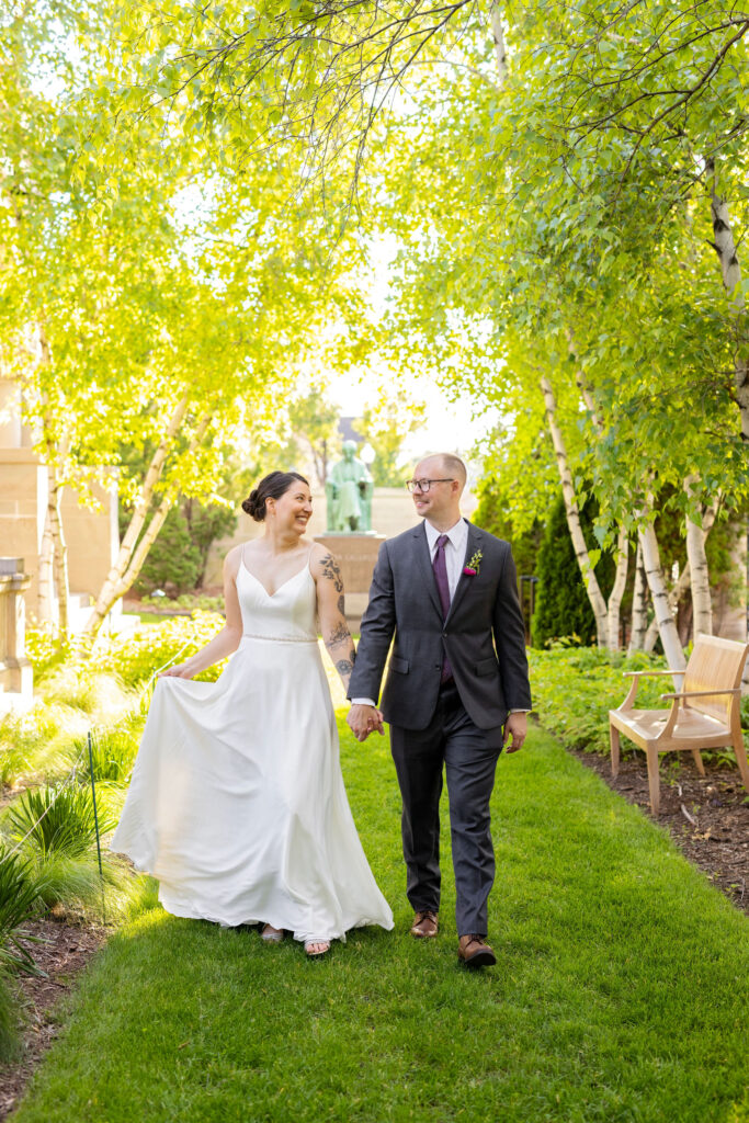 Bride and groom walk hand-in-hand through a sunlit garden, smiling and enjoying a quiet moment together.