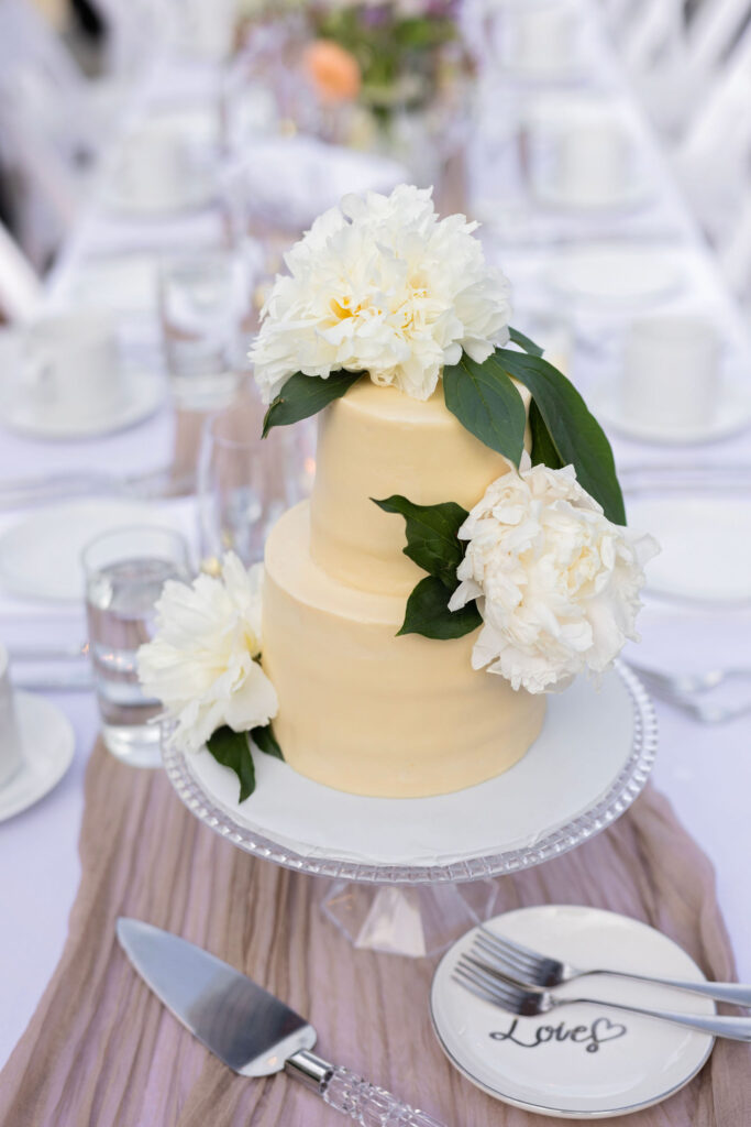 Three-tiered ivory wedding cake adorned with fresh white flowers and displayed on a glass cake stand at the center of a reception table.