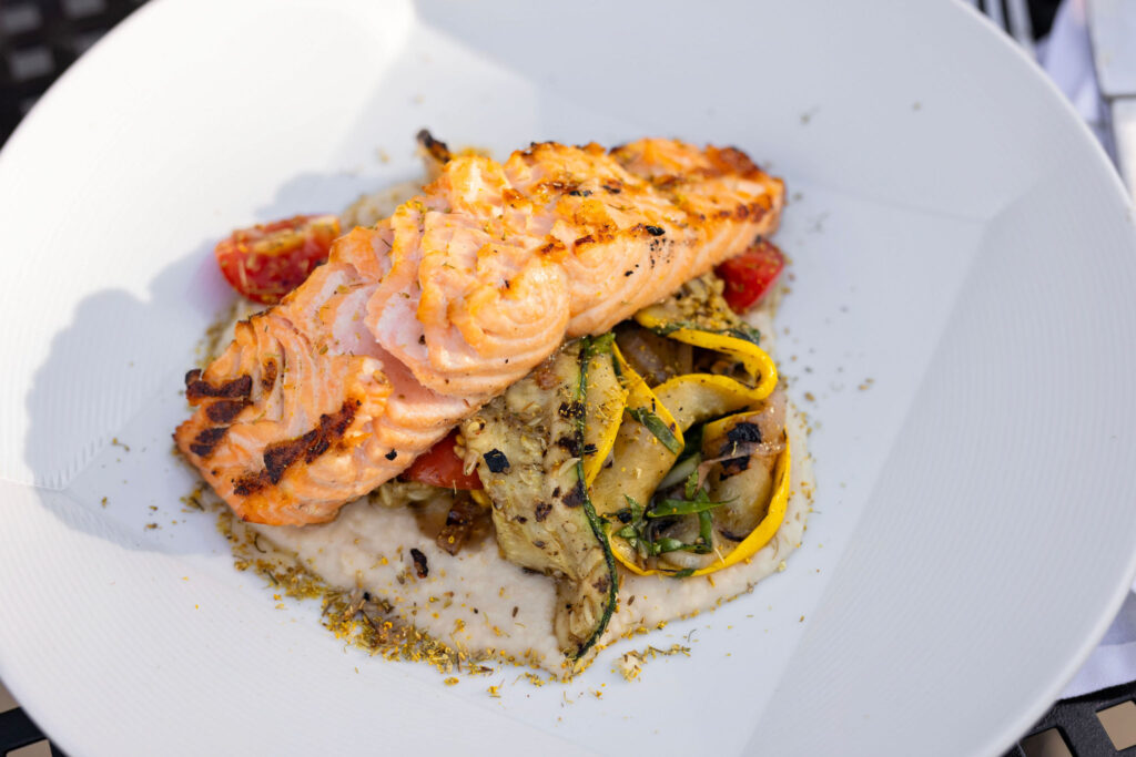 Grilled salmon served over creamy polenta with roasted summer vegetables including zucchini and cherry tomatoes.