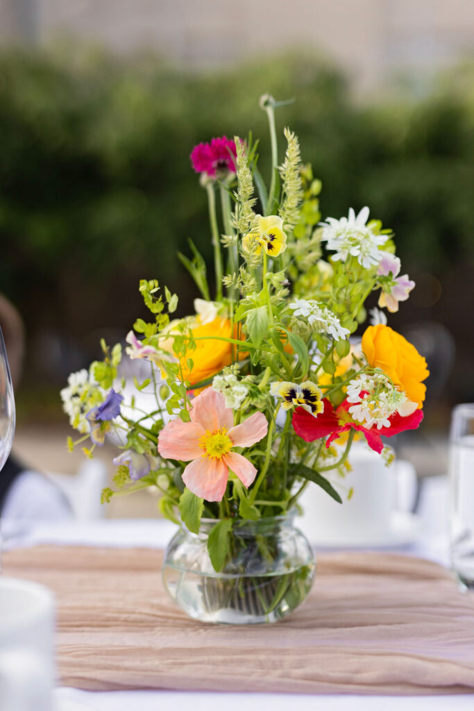 A vibrant, garden-style floral centerpiece with daisies, poppies, and wildflowers in a small glass vase on a white-clothed table.