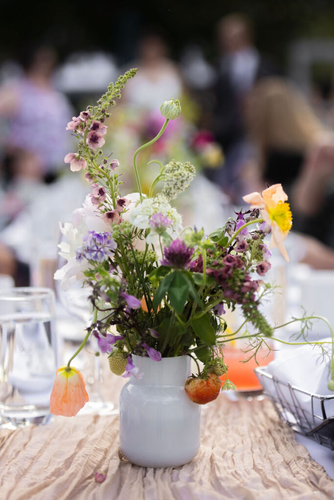 Close-up of a wildflower centerpiece in a ceramic white pitcher, surrounded by glassware and soft beige table runners.